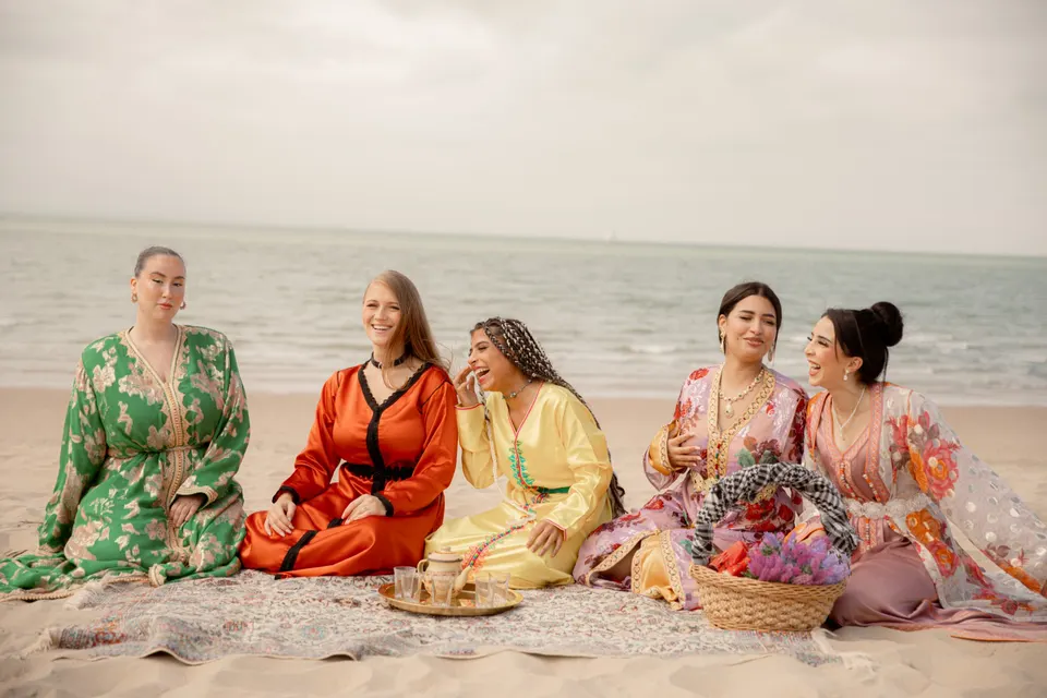 Cinq femmes en caftans colorés rient, assises sur une plage autour d’un plateau à thé, photographie de Women in Black Photography
